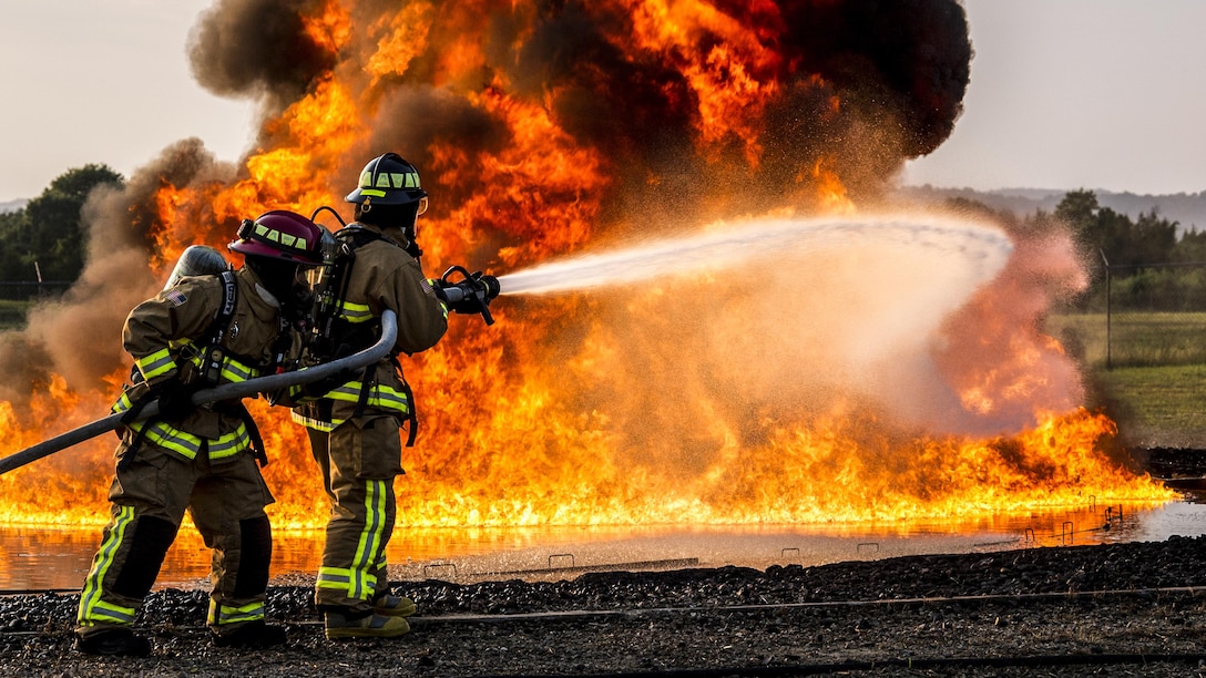 Two airmen fight a large flame with water from a hose.