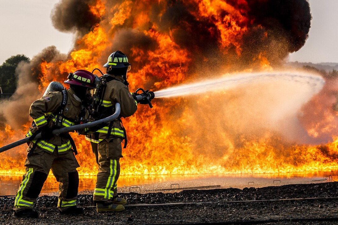 Two airmen fight a large flame with water from a hose.