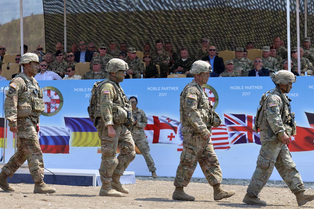 Soldiers walk in front of a crowd of people in stands.