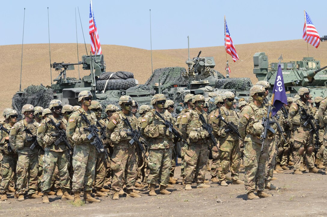 Soldiers stand in formation in front of military vehicles with American flags.