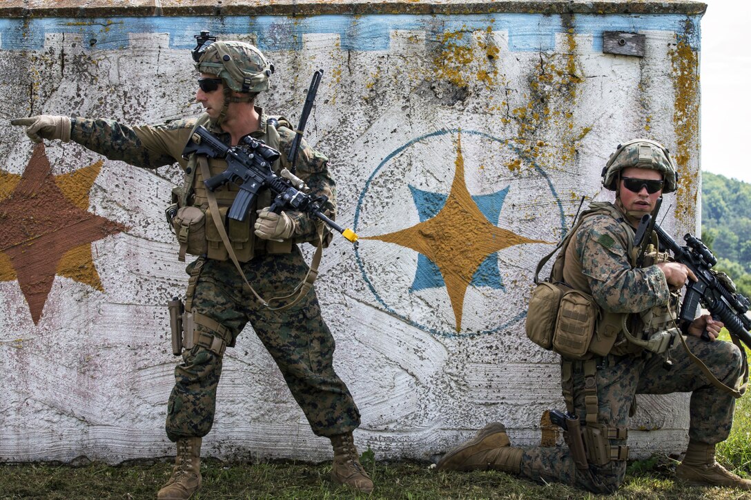 One Marine kneels by a wall while another stands and points.