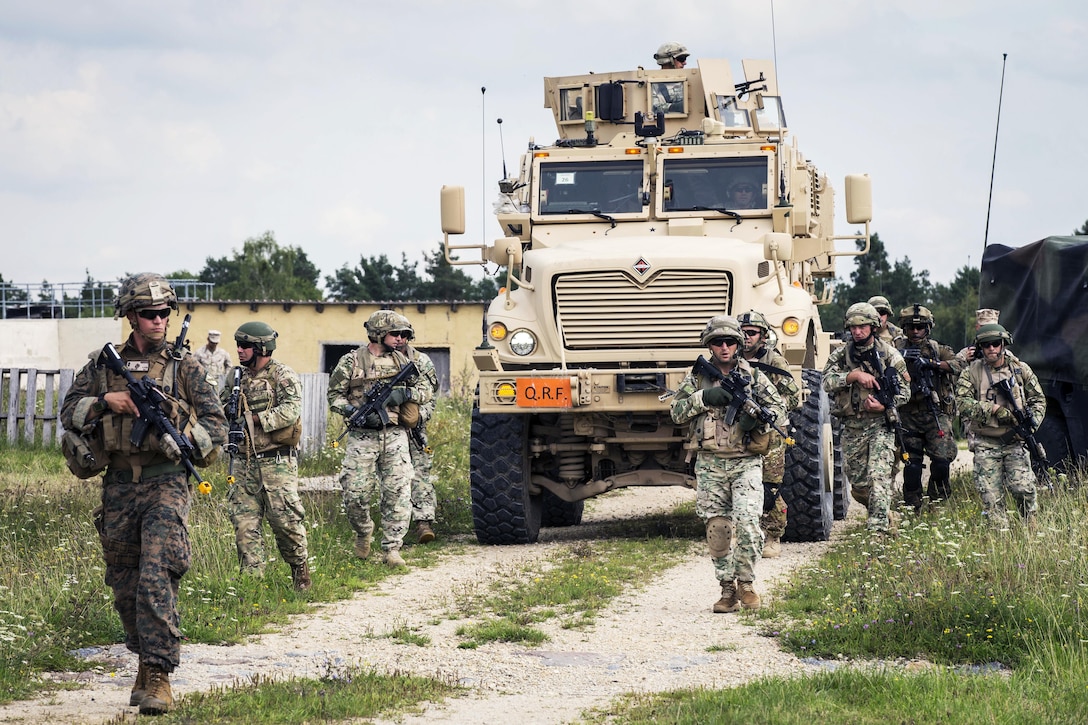 Marines carry weapons and walk in a loose formation in front of a tactical vehicle.