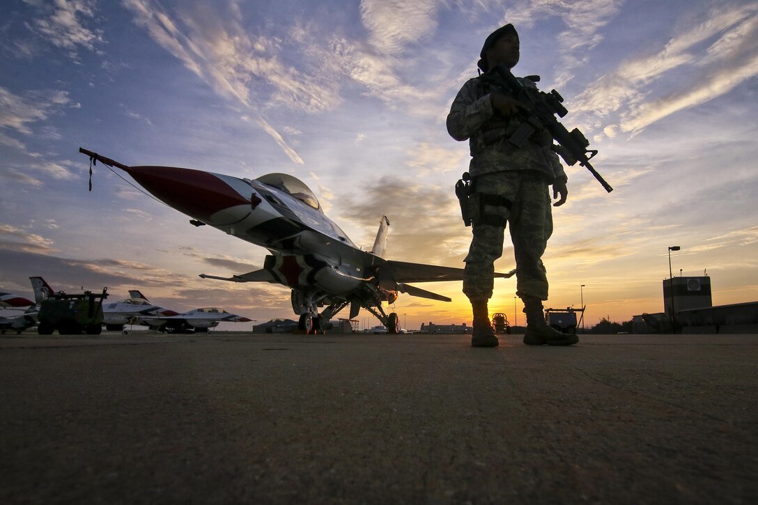 An airman stands with her weapon next to an aircraft on a flightline at sunrise.
