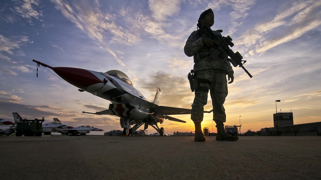 An airman stands with her weapon next to an aircraft on a flightline at sunrise.