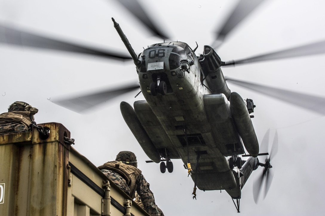 A helicopter descends toward a Marine crouching on top of a metal container.