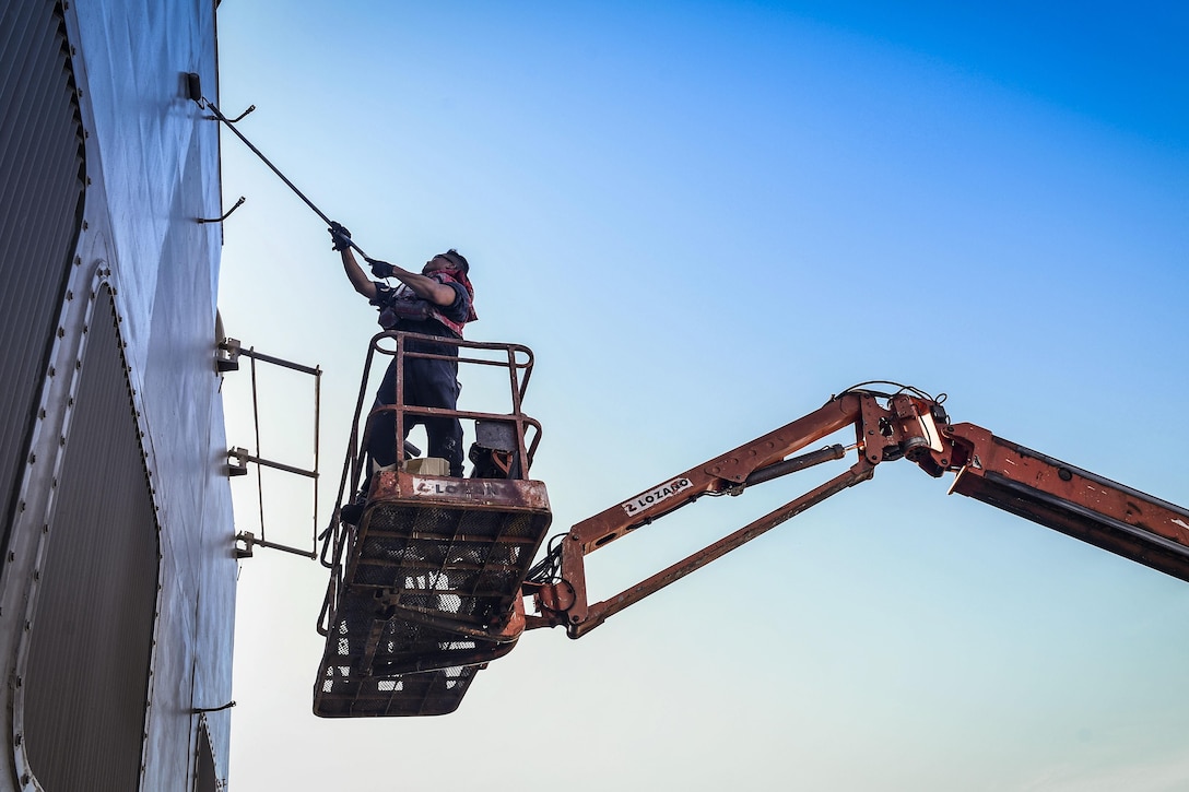 A sailor on elevated scaffolding uses a roller brush to paint the side of a ship.