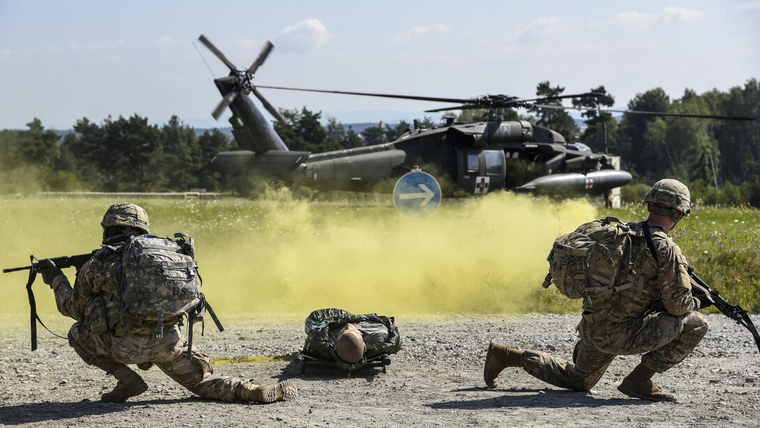 Two soldiers wield weapons with a mannequin on the ground between them and a helicopter nearby, behind a yellow smoke cloud.