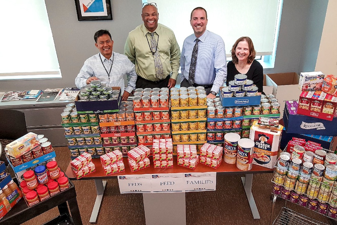 Four people stand behind a table display of nonperishable food items.