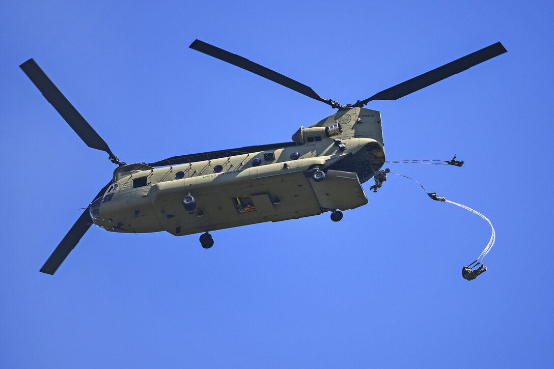 Soldiers jump out of the back of a helicopter in front of a bright blue sky.