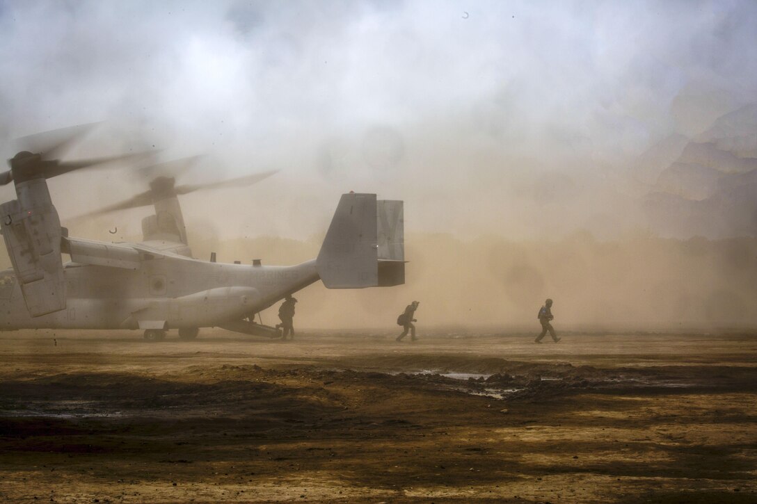 Troops move out from the back of an aircraft as dust billows in the background.