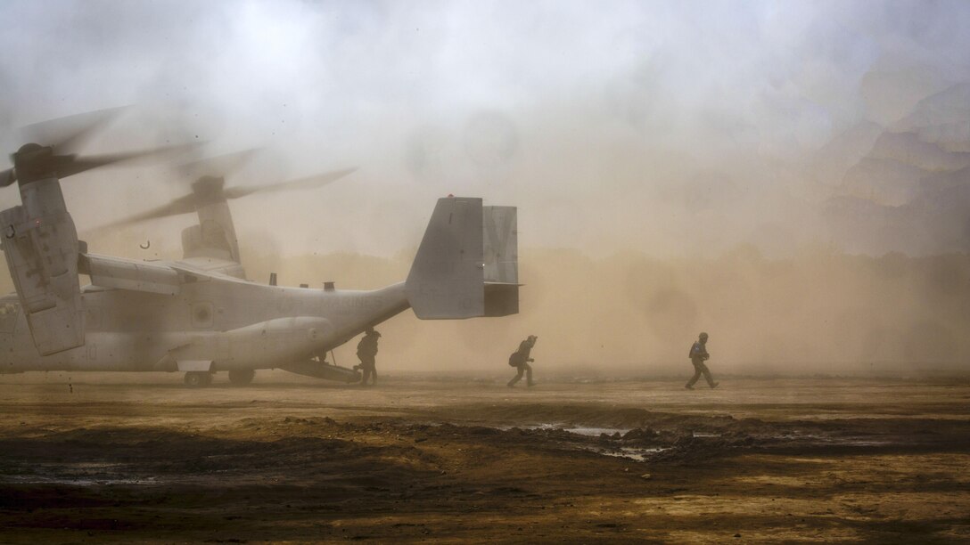 Troops move out from the back of an aircraft as dust billows in the background.