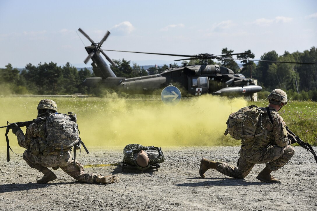 Two soldiers wield weapons with a mannequin on the ground between them and a helicopter nearby, behind a yellow smoke cloud.