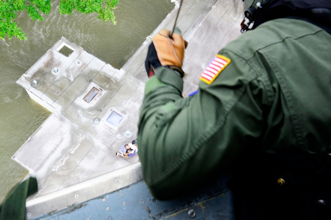 A Coast Guardsman lowers a cable toward the roof of a building surrounded by water.