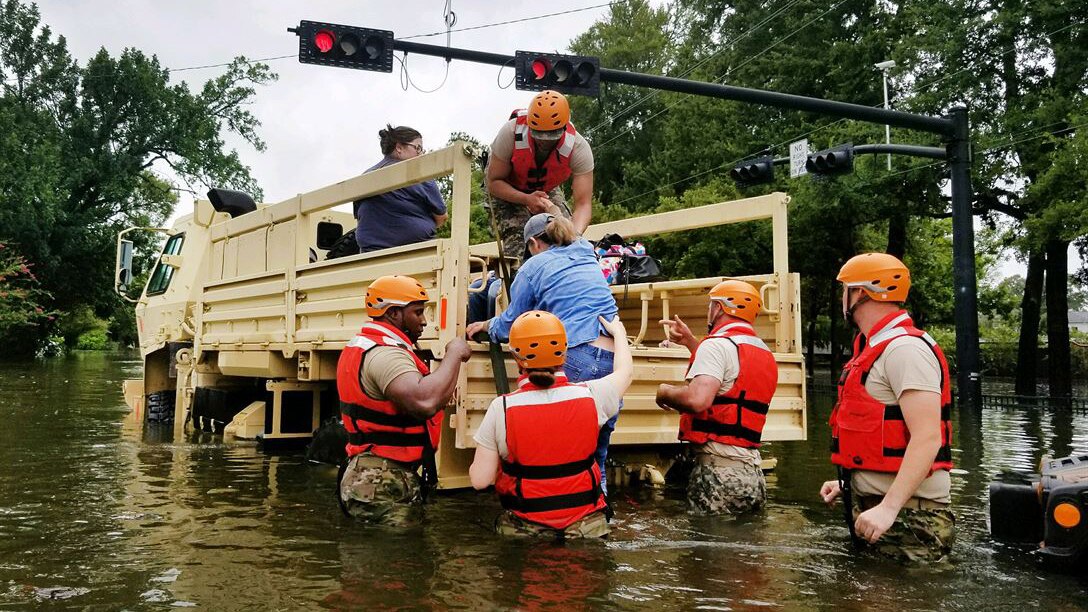 Several guardsmen wearing helmets and life vests and standing in waist deep water are helping woman onto the back of a military vehicle.