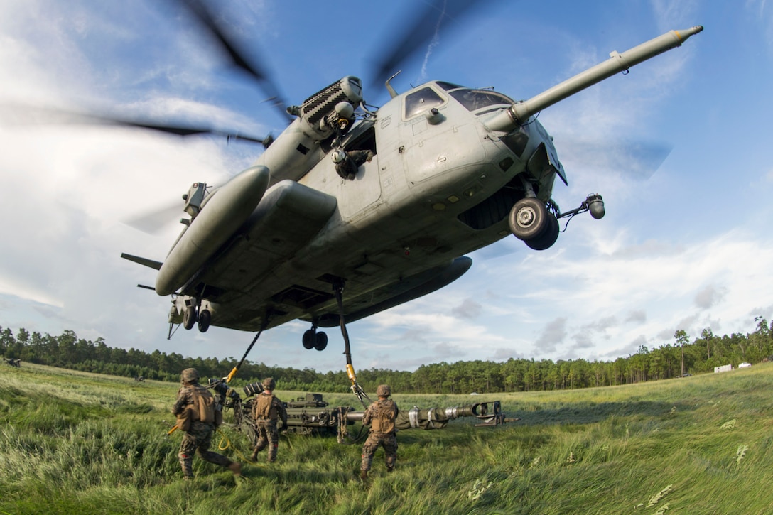 A helicopter hovers over Marines as they work to attach a howitzer to it by sling load.