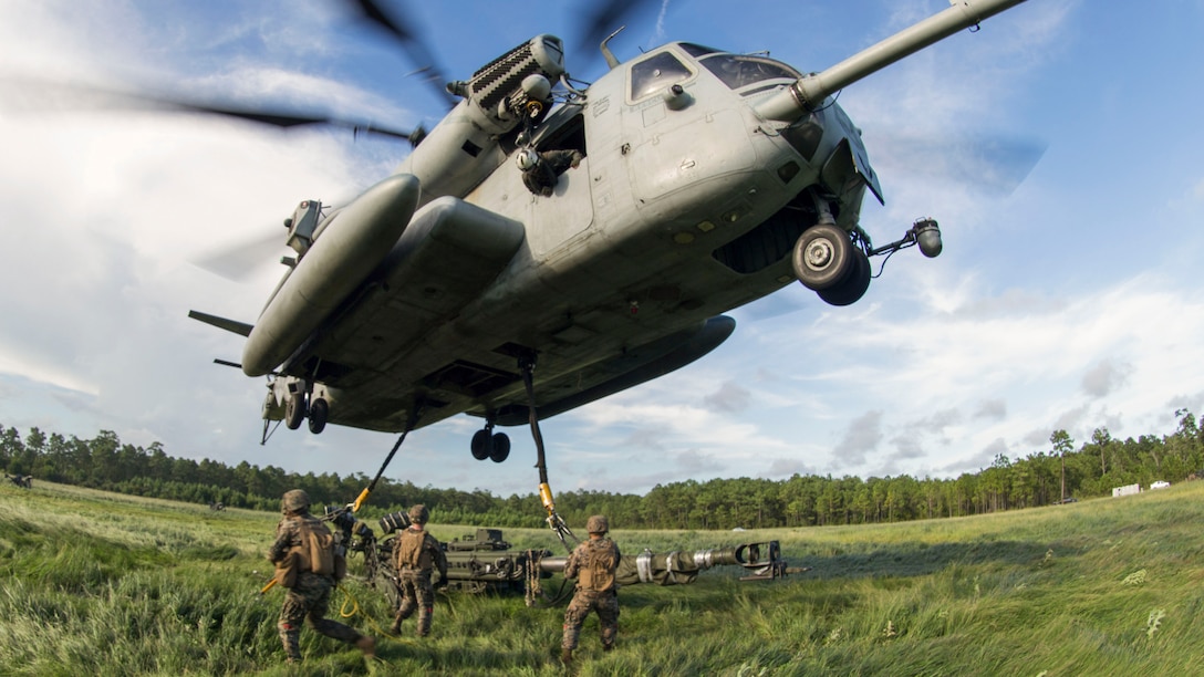 A helicopter hovers over Marines as they work to attach a howitzer to it by sling load.