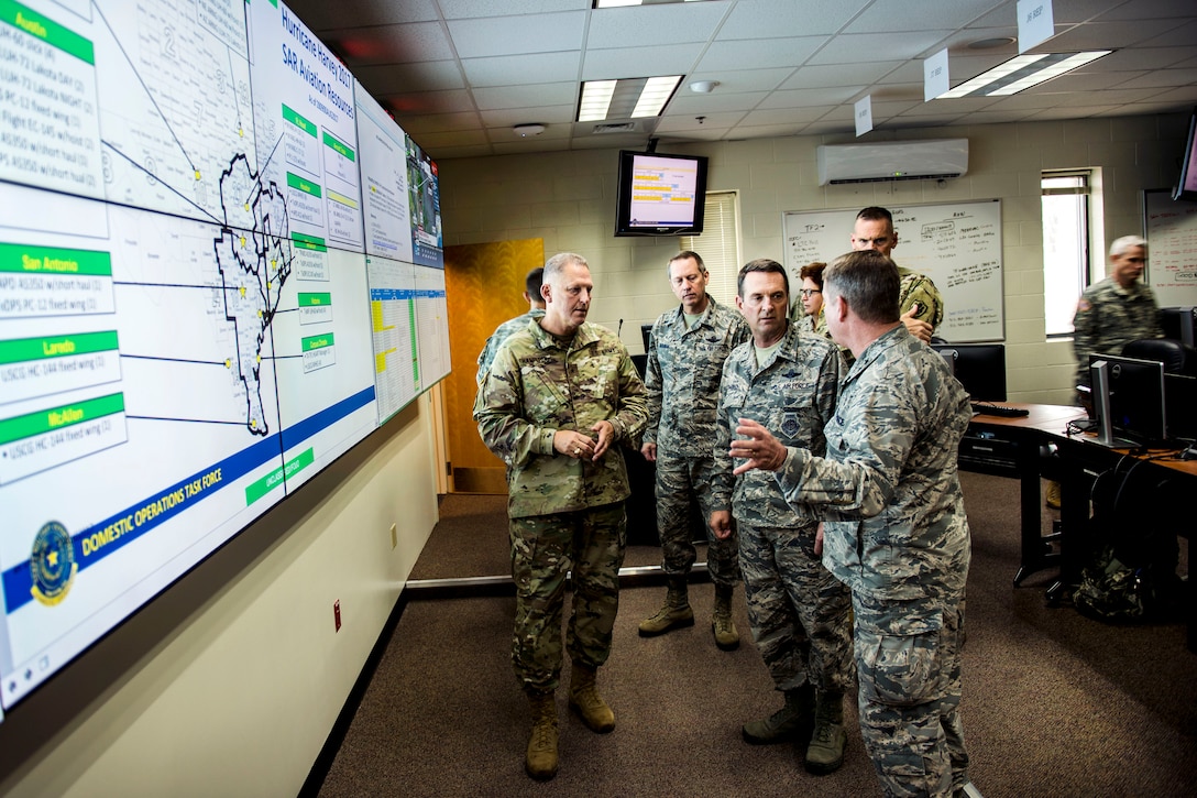 A group of airmen and soldiers talk near of a large map on a wall.