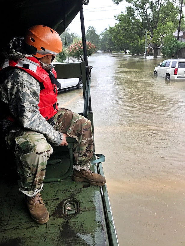 A guardsman sits in the back of a military vehicle and looks out over flooding streets.
