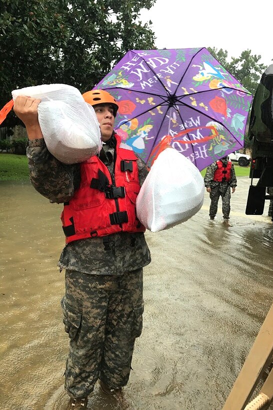 A guardsman holds items and an umbrella while standing in a flooded street.