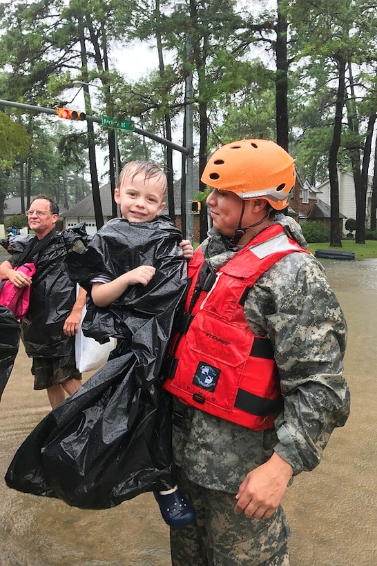 A guardsman holds a small child while standing in a flooded street.