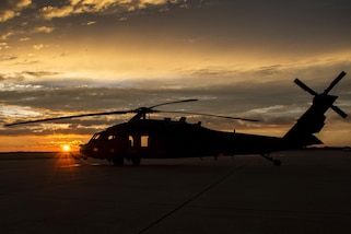 A helicopter sits on the ground with the sun behind it.