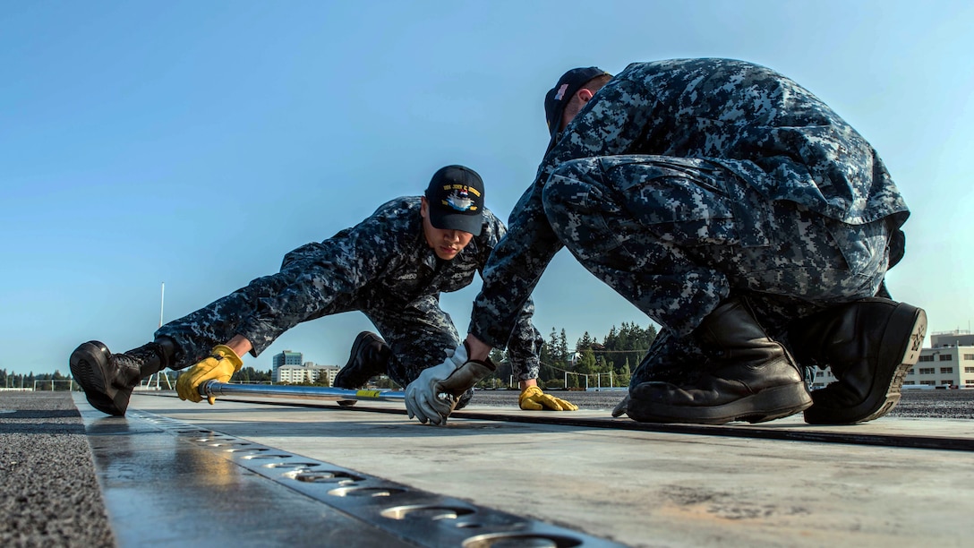 Two sailors kneel on a flight deck while doing maintenance work.