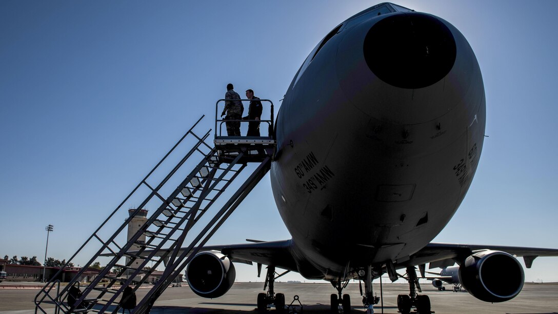 Silhouettes of two airmen stand on a platform on top of a movable staircase by the door of an aircraft.