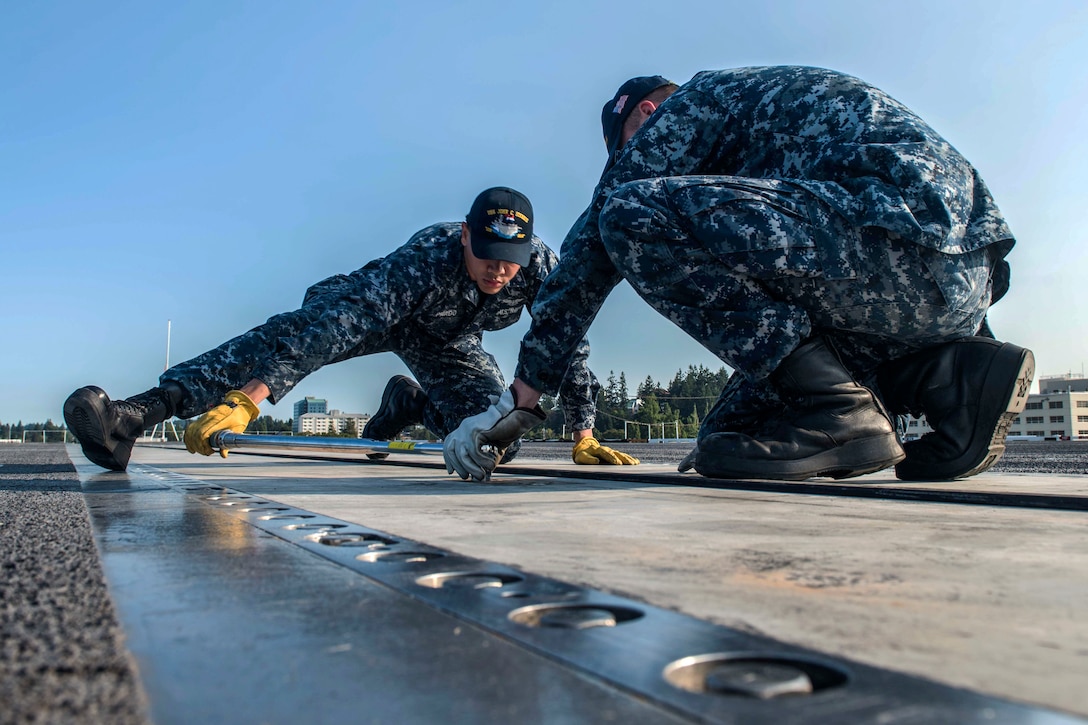 Two sailors kneel on a flight deck while doing maintenance work.