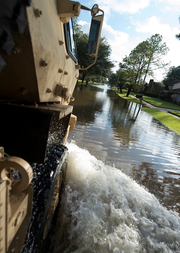 A military vehicle drives through water in a flooded street.
