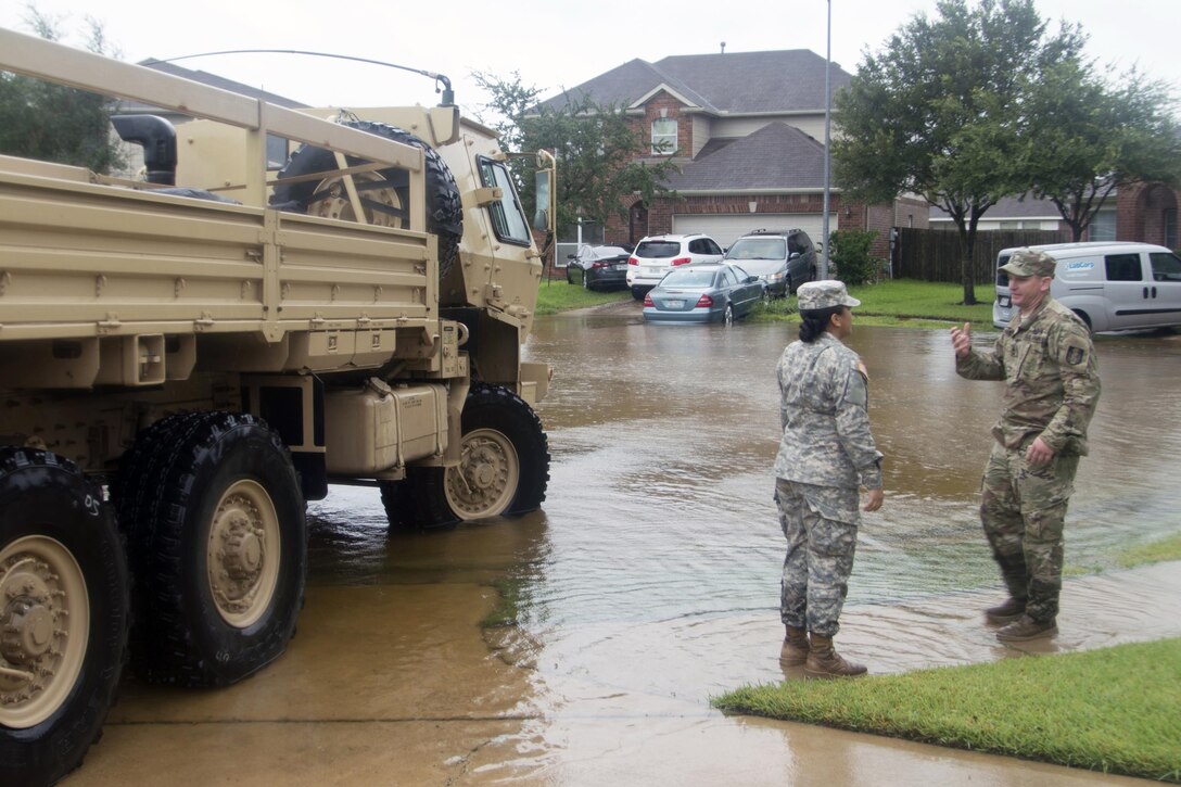 Two people stand next to a military vehicle in a flooded street.