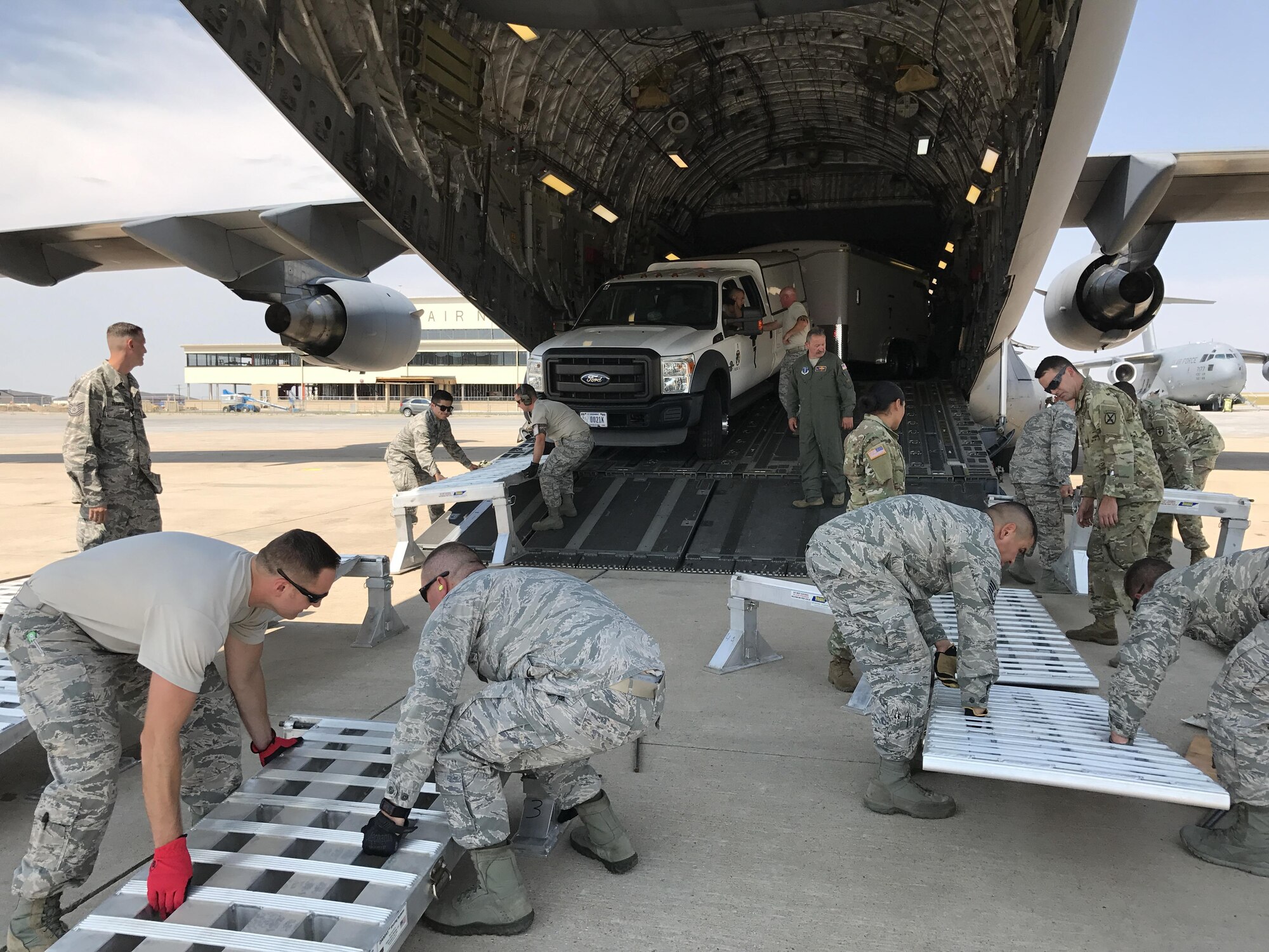 Airmen work together to move pieces of a ramp, while a truck and trailer are backed into a C-17 aircraft behind them