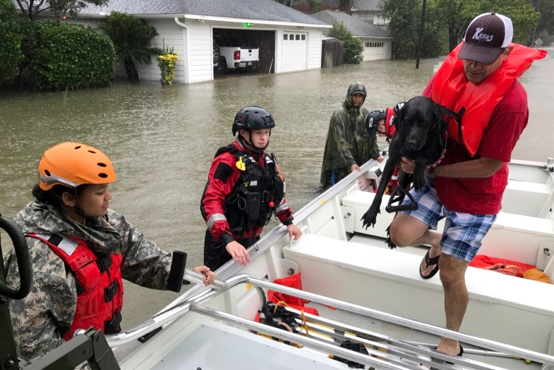 Three people stand in water and hold a boat steady while a person holding a dog walks in the boat.