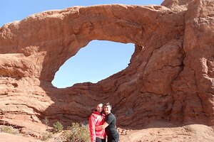 A man hugs a woman standing in front of "The Windows" natural rock feature at Arches National Park.