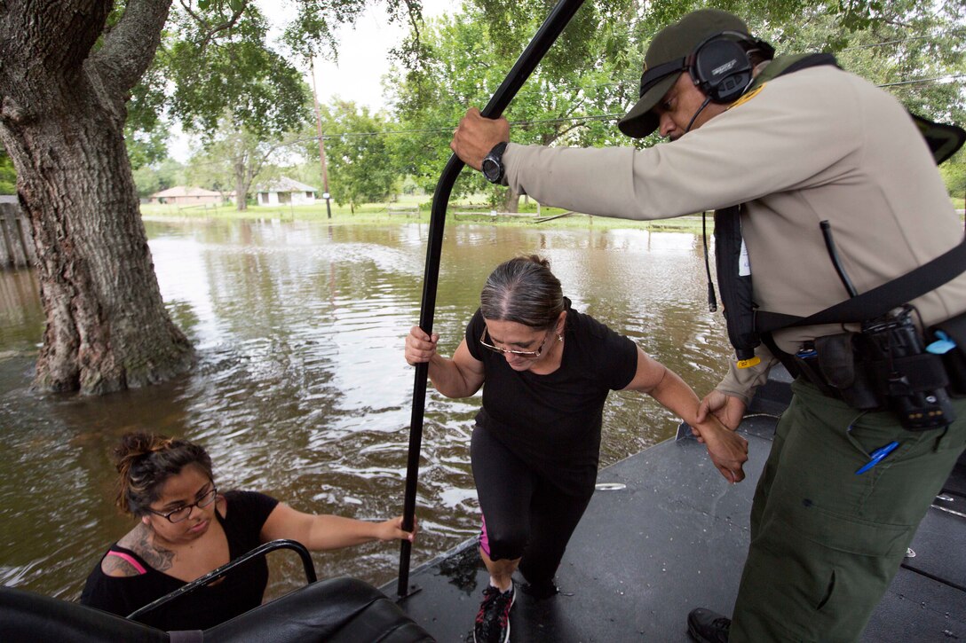 A Border Patrol agent helps a woman board a boat as another woman stands in chest-deep waters preparing to board.