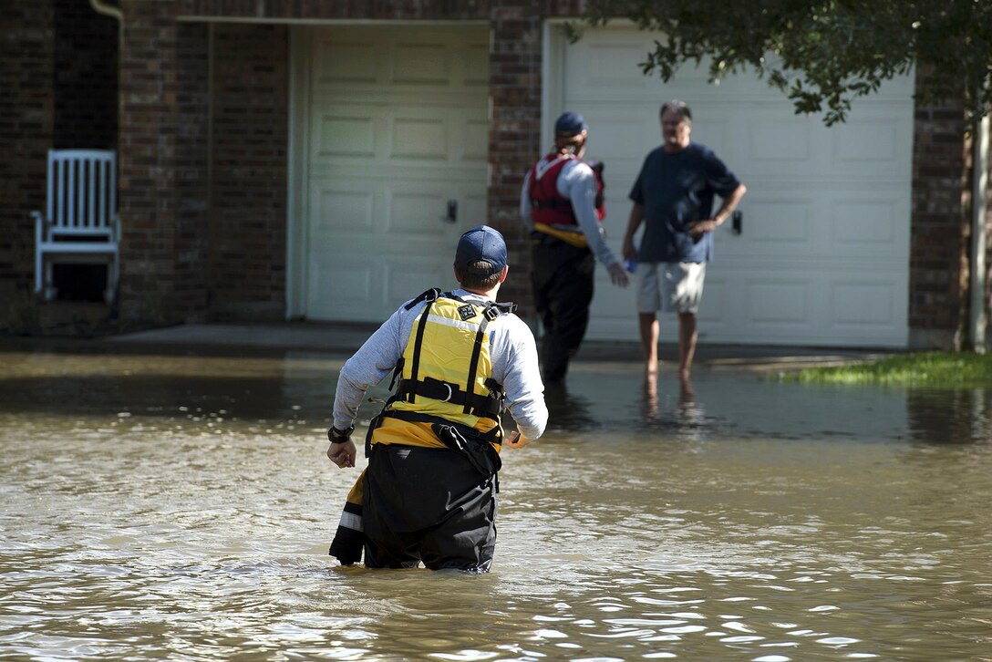 A man standing in waist-deep water looks towards a house where two people are standing in ankle-deep water.