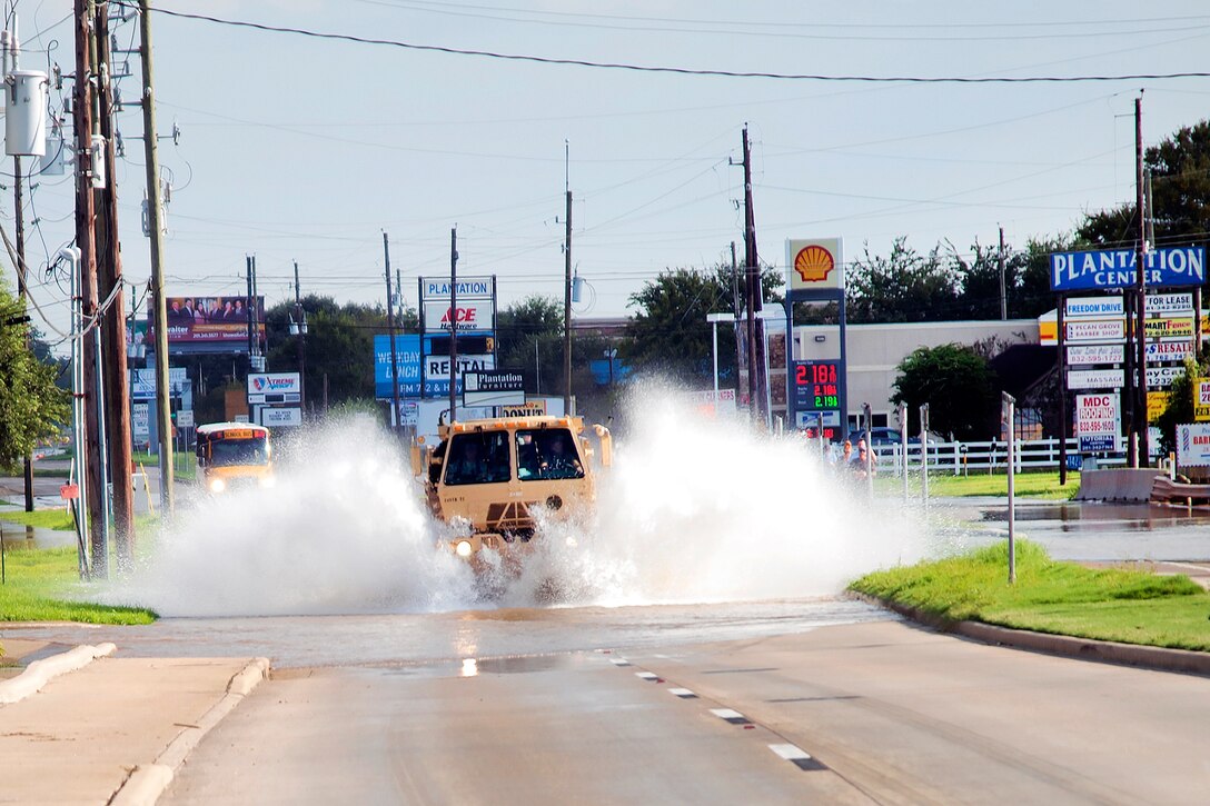 A military tactical vehicle creates a splash as it drives on a flooded street.