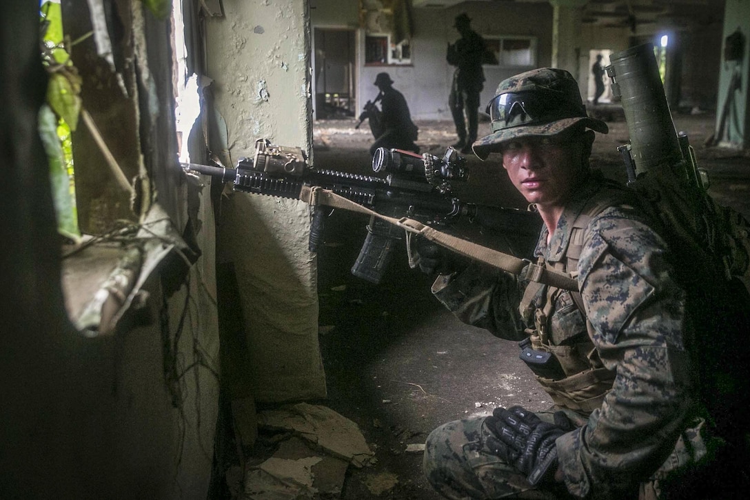 A Marine holds a weapon and looks around for threats inside a building with other Marines.