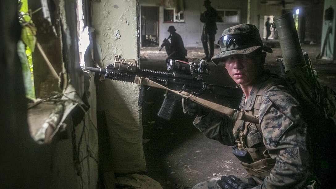 A Marine holds a weapon and looks around for threats inside a building with other Marines.