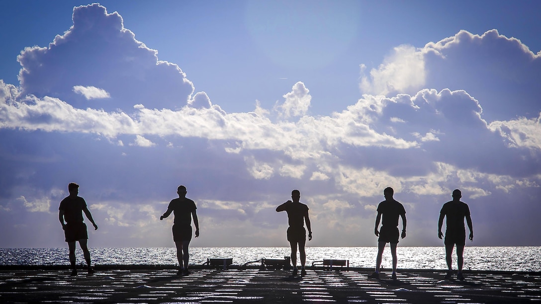Five Marines, shown in silhouette, stand on the deck of a ship in the ocean.