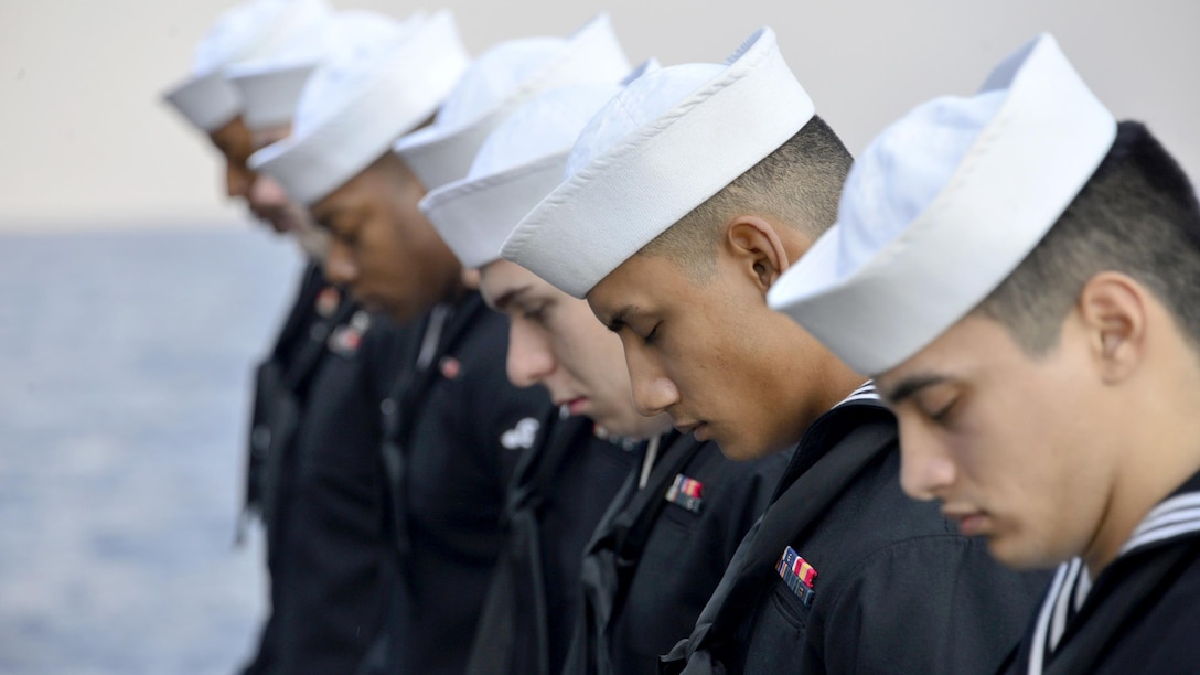 A row of sailors bow their heads with eyes closed, with the sea as a backdrop.