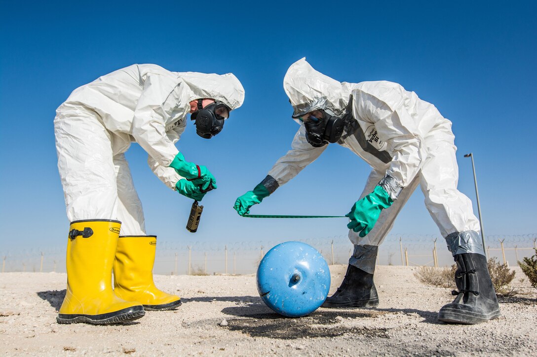 Two airmen in white protective body suits and gloves lean over a simulated explosive in the desert.