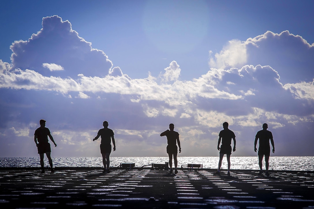 Five Marines, shown in silhouette, stand on the deck of a ship in the ocean.