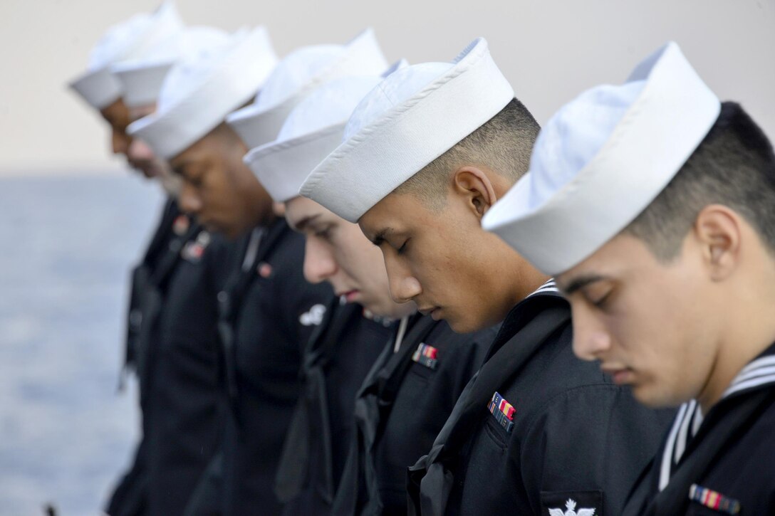 A row of sailors bow their heads with eyes closed, with the sea as a backdrop.