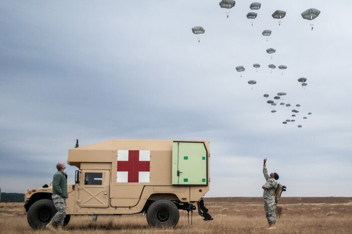 Soldiers standing by a military ambulance look up at descending parachutists in a drop zone.
