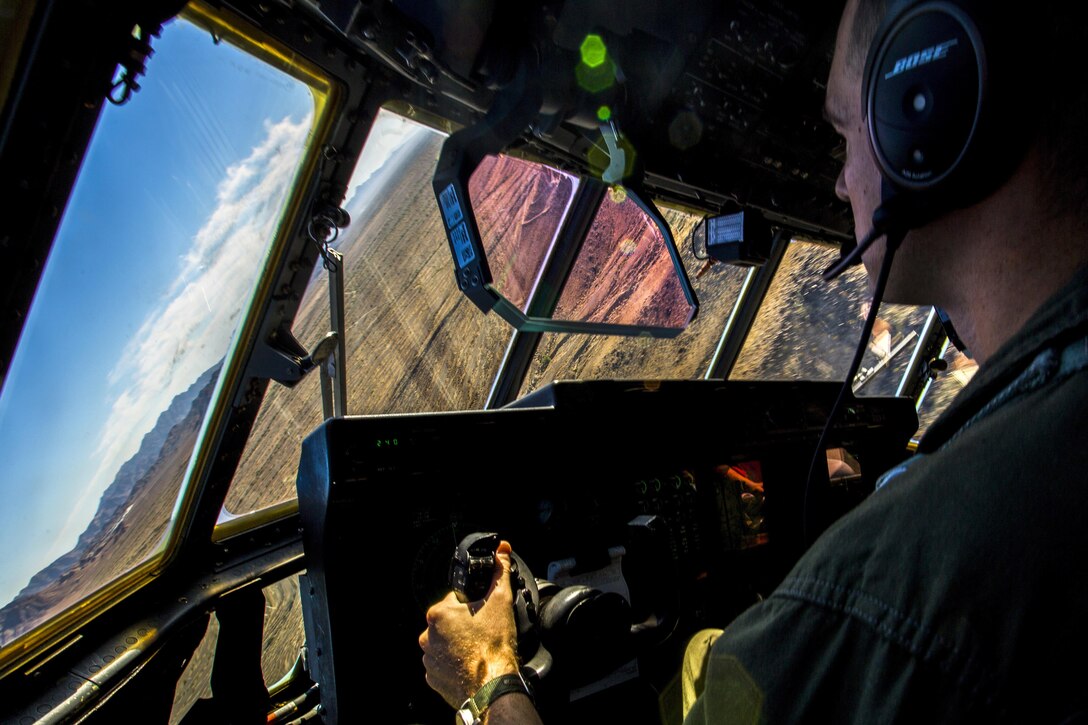 A pilot looks out a cockpit window at an angled view of dessert terrain and blue sky.
