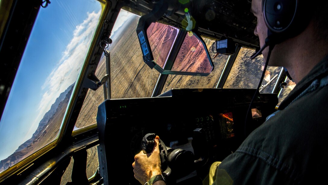 A pilot looks out a cockpit window at an angled view of dessert terrain and blue sky.