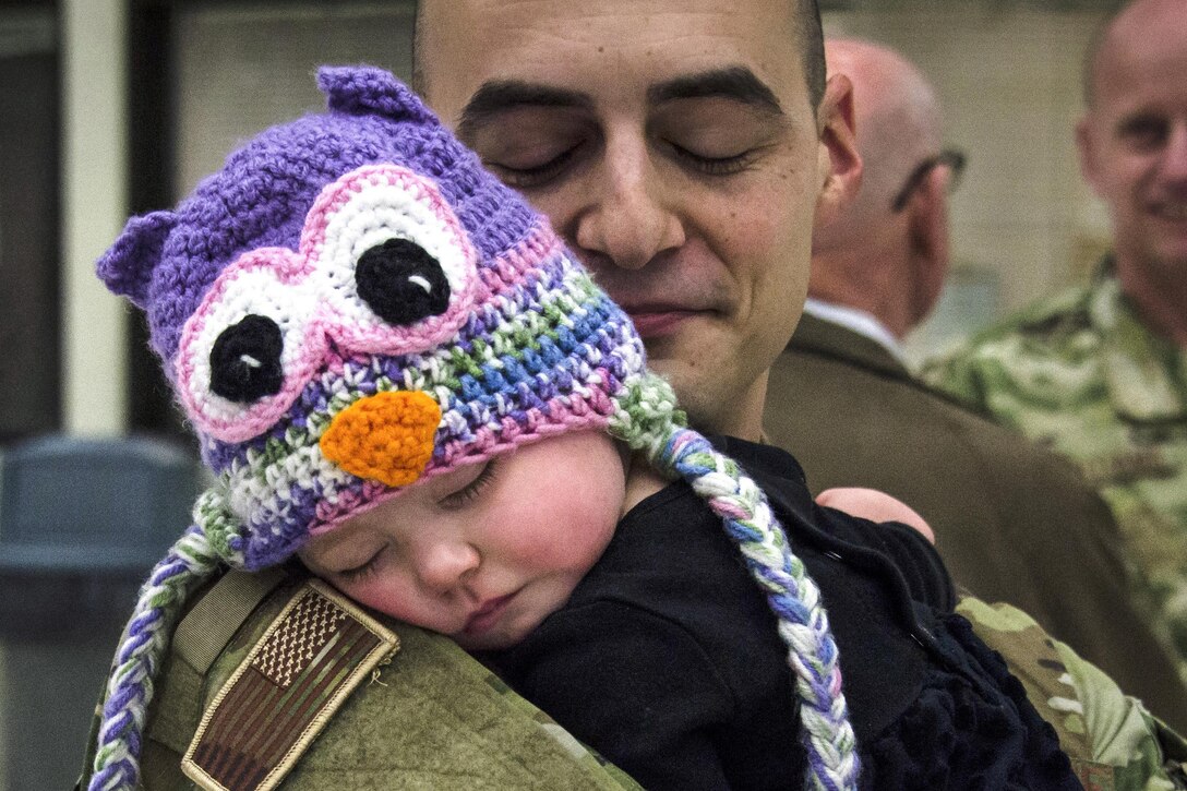 An airman holds a sleeping baby wearing a knitted cap with an owl's face.