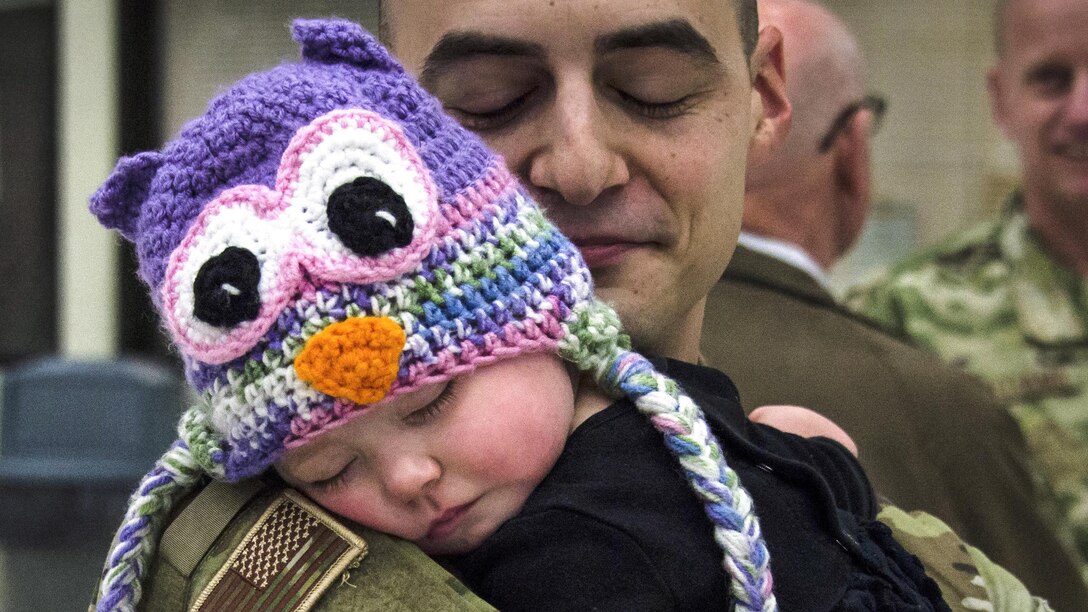An airman holds a sleeping baby wearing a knitted cap with an owl's face.