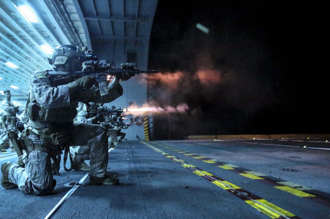 Marines, illuminated by blue light, kneel and fire weapons on a ship's flight deck.