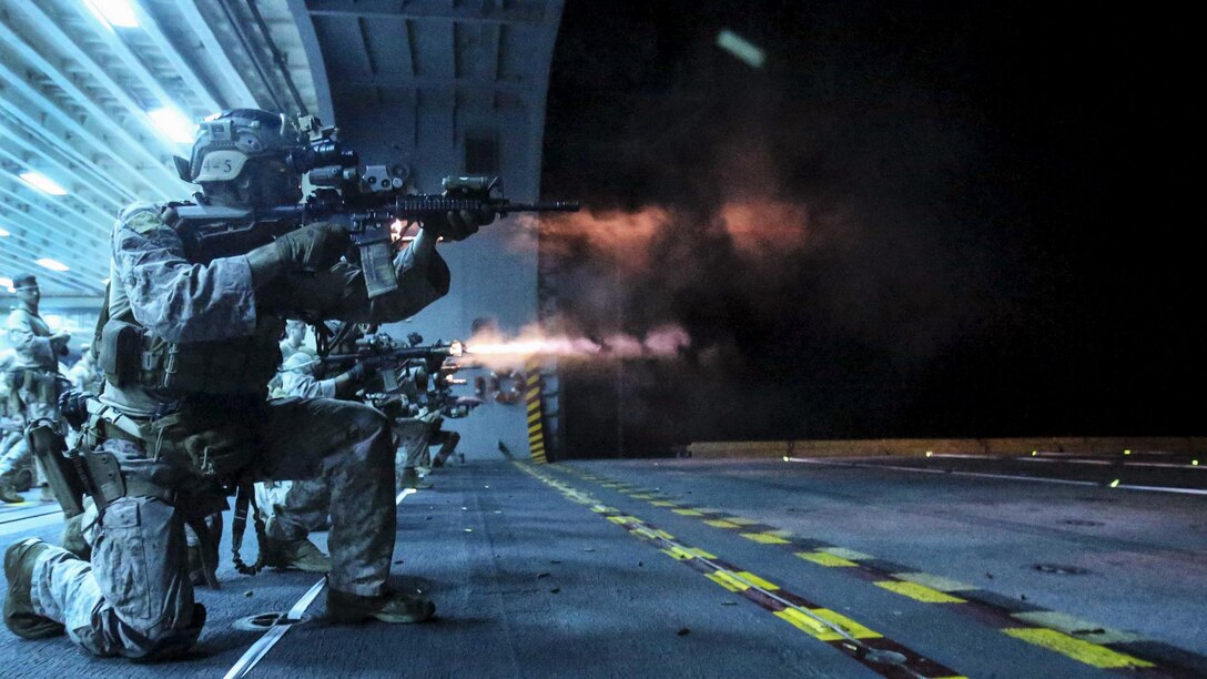 Marines, illuminated by blue light, kneel and fire weapons on a ship's flight deck.